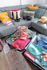 Close up of open suitcase with summer clothes and accessories placed on coffee table and sofa in living room, symbolizing upcoming vacation and travel preparations