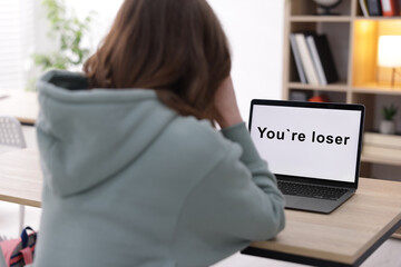 Cyber bullying. Upset teenage girl and laptop with text You Loser at desk indoors