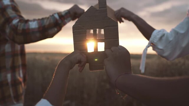 Sun shining through cardboard symbol of home, family walking in field in sunrise . Refugees dreaming about calmness and happiness, need own house and peaceful life, move to dream, parents and children