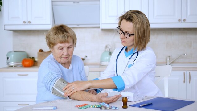 Young female doctor is measuring blood pressure of a senior woman during home visit, pills and digital blood pressure monitor are on the table