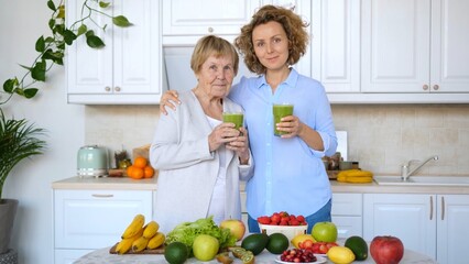 Senior woman and her daughter are embracing and holding glasses of green smoothies in a kitchen full of fresh fruits and vegetables, promoting healthy eating habits and intergenerational bonding