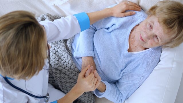 Young female doctor in a white coat checking on a senior patient lying in bed, holding her hand gently and providing comfort and support during a difficult time