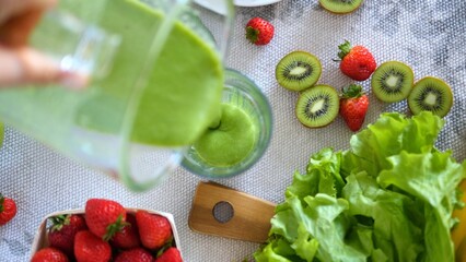 Chef pours a vibrant green smoothie from a blender into a glass, surrounded by fresh strawberries, kiwi slices, and lettuce, showcasing a healthy and refreshing culinary creation