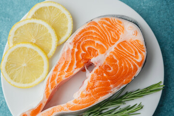 Closeup raw salmon steak with lemon slices and rosemary on white plate over blue background