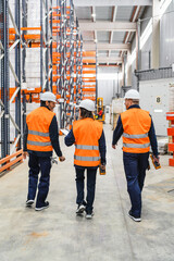 Warehouse workers wearing safety vests and helmets are walking through large distribution center, using barcode scanner and discussing logistics operations