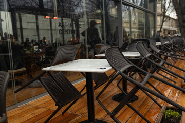 An empty outdoor seating area of a cafe with chairs placed upside down on tables, likely closed due to cold weather. Reflections in the glass show a quiet city street and indoor patrons.