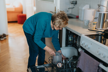 An elderly woman actively managing her household chores, focusing on arranging dishes in the kitchen dishwasher.