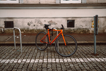Bright orange bicycle is parked and secured to a metal bike rack on a cobblestone street. Behind it stands a historic beige building with white-framed windows, evoking a calm, urban European setting