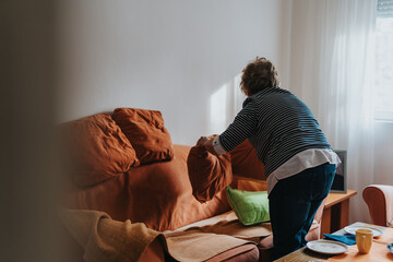 An elderly woman arranging cushions on a couch, creating a cozy atmosphere in a room with natural...