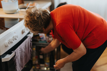 A senior woman wearing a red blouse is bending to open a kitchen drawer, highlighting an everyday and relatable moment in a comfortable and warm domestic setting.