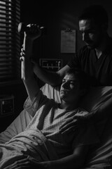 A boy lifts a dumbbell in bed with support from a therapist, captured in dramatic black and white light and shadow contrast.