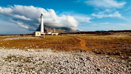 Lighthouse on italian coast of Sicily with path on yellow field and blue sky with white clouds. Bautiful landscape of nature. Travel on island.