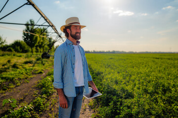 Farmer with a straw hat and a beard is holding a tablet and looking at his crops in a cultivated...