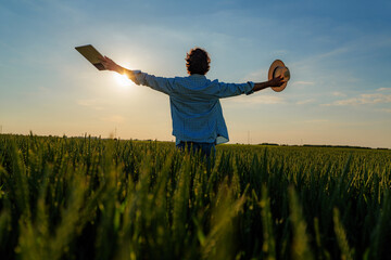 Farmer stands in a wheat field at sunset, holding a tablet in one hand and a straw hat in the...