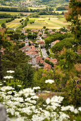 Picturesque View Of Vipava From Castle Ruins: Red-Tiled Rooftops And Church Tower Nestled In Green Valley With Blooming Flowers In Foreground And Rolling Fields In Background, Slovenia