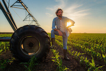 Confident farmer leaning against a tractor wheel, holding his hat while watching over a corn field...