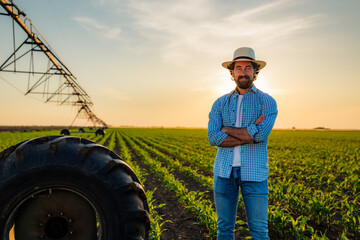 Confident farmer standing proudly with crossed arms in his corn field at sunset, an irrigation...
