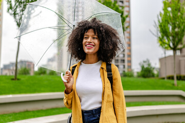 Young woman smiling under an umbrella while enjoying a rainy day in a city park with green grass...