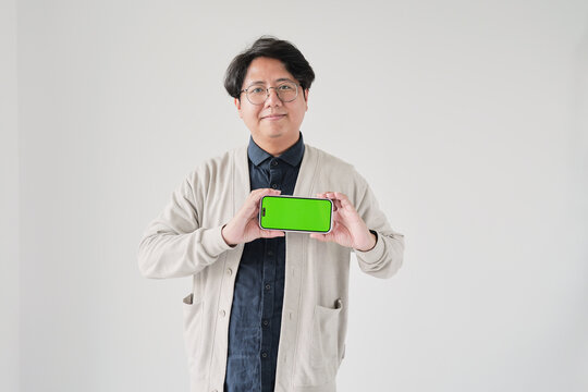 A man is holding a green phone. He is wearing glasses and a white sweater. The phone is green and has a green screen - Powered by Adobe