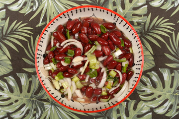 A vibrant bean salad with red kidney beans, sliced white onion, and green onions is presented in a patterned bowl on a tropical leaf-patterned tablecloth