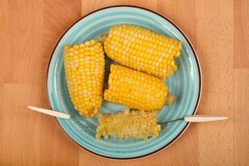 Three yellow corn cobs and one eaten cob with corn holders are arranged on a blue plate, viewed from above on a wooden surface