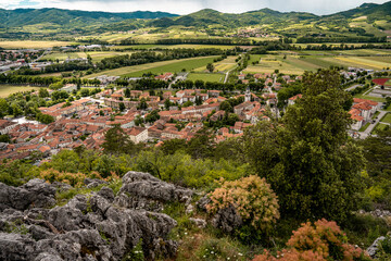 Picturesque View Of Vipava From Castle Ruins: Red-Tiled Rooftops And Church Tower Nestled In Green Valley With Blooming Flowers In Foreground And Rolling Fields In Background, Slovenia