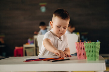 Cute boy is sitting by the table and playing. Group of children are in the kindergarten