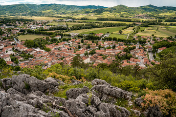 Picturesque View Of Vipava From Castle Ruins: Red-Tiled Rooftops And Church Tower Nestled In Green Valley With Blooming Flowers In Foreground And Rolling Fields In Background, Slovenia