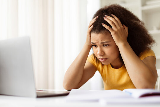 Anxious black woman touching her head while looking at laptop screen, upset stressed african american female freelancer sitting at desk in her home office, suffering problems with work