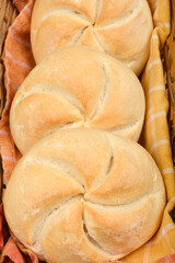 A close-up, top-down view shows three freshly baked Kaiser rolls nestled in a woven basket on a...