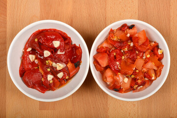 Two white bowls on a wooden background: the left one holds a roasted pepper and garlic salad, and the right one contains a roasted pepper, tomato, and garlic salad
