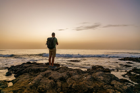 Solo traveler with backpack standing on rocky coastline watching golden sunset over Atlantic ocean waves during journey in Tenerife