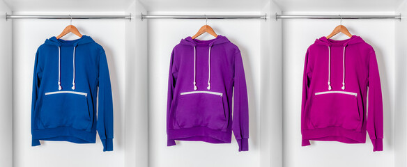 Three colorful hoodies in blue, purple, and magenta hanging on wooden hangers inside white...