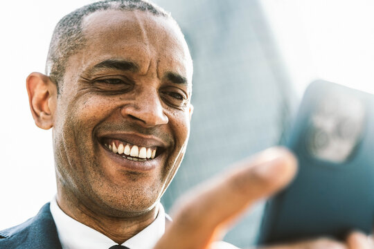 Smiling black businessman holding smart mobile phone in front of office building - Happy business man using online banking app on smartphone - Technology and business life style concept