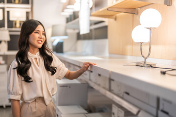 Asian woman testing decorative lamp at home furniture store touching light fixture to evaluate...