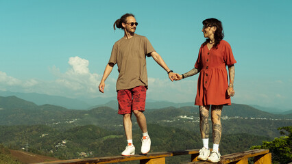 Couple holding hands and balancing on a wooden beam with mountains in the background. Smiling,...
