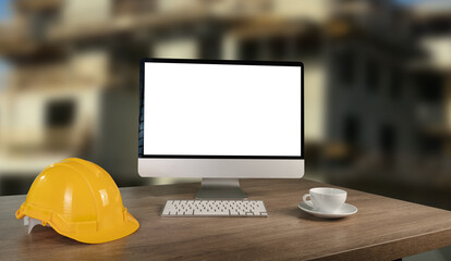 A laptop is open on a desk with a yellow hard hat and a red cup of pens. The laptop is empty and the desk is cluttered with construction tools. working table architect designer