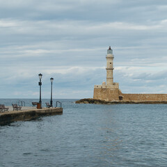 Fototapeta premium Old Venetian lighthouse in Chania, Crete, Greece. Seaside view with calm waters, stone pier, benches, and cloudy sky over the Mediterranean