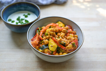 Vegetarian curry made from chickpeas and vegetables such as zucchini, peppers and leek in a bowl, served with fresh yogurt, healthy meal on a wooden table, copy space