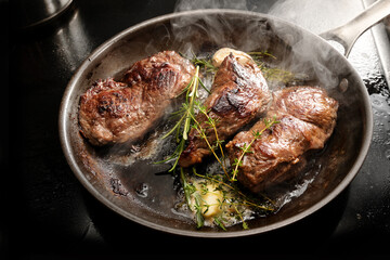 Fresh steaming beef steaks are seared in a carbon steel frying pan with butter, garlic, thyme and rosemary, cooking tasty meat, selected focus