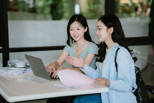 Distant learning. Two smiling diverse trendy teenage girls college university students use laptop for online studying at library take notes from video lesson prepare task surf information at websites