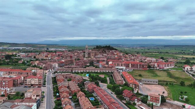 Smooth aerial orbit around Navarrete, La Rioja. Scenic view of historic town, church tower, red rooftops, vineyards and green countryside under cloudy sky