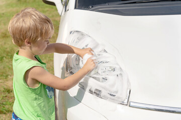 A boy in a green T-shirt rubs the headlights with a sponge and foam. He is washing the car.
