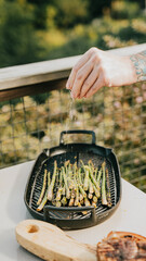 A person sprinkling salt on a tray of asparagus. The tray is black and the asparagus are green