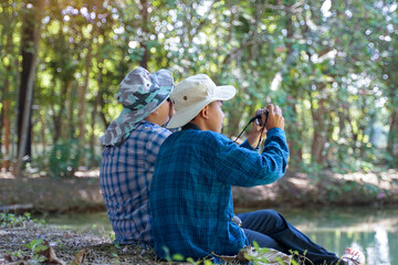 Two Asian boys use binoculars and a camera to study the local birds in a community forest. Soft and selective focus.
