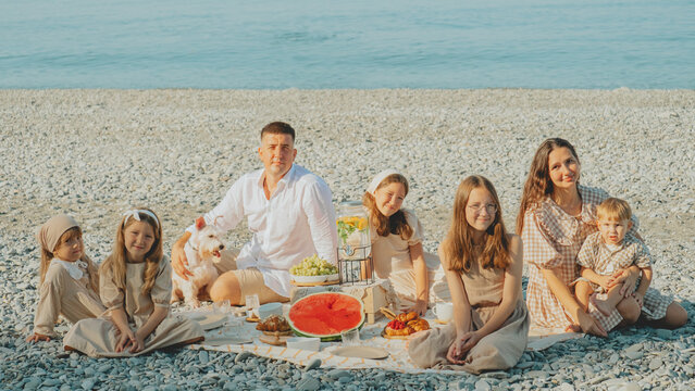 A big family enjoys a summer picnic on a pebble beach by the sea — kids, parents, and a dog spending a sunny day together with fruits, pastries, and refreshing drinks.