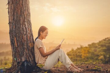 A student girl with glasses sitting under a pine tree reading book sunset background.