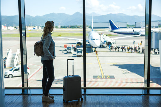 Woman with suitcase waiting at airport terminal before boarding airplane, solo travel passenger ready for flying to summer vacation - Powered by Adobe