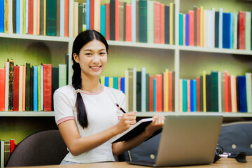 An Asian female university student sits in the library with her laptop, happily researching and working on academic tasks in a calm and focused environment.