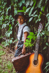 boy dressed in the style of the 20s stands next to a guitar holding a suitcase in his hand looking at the camera